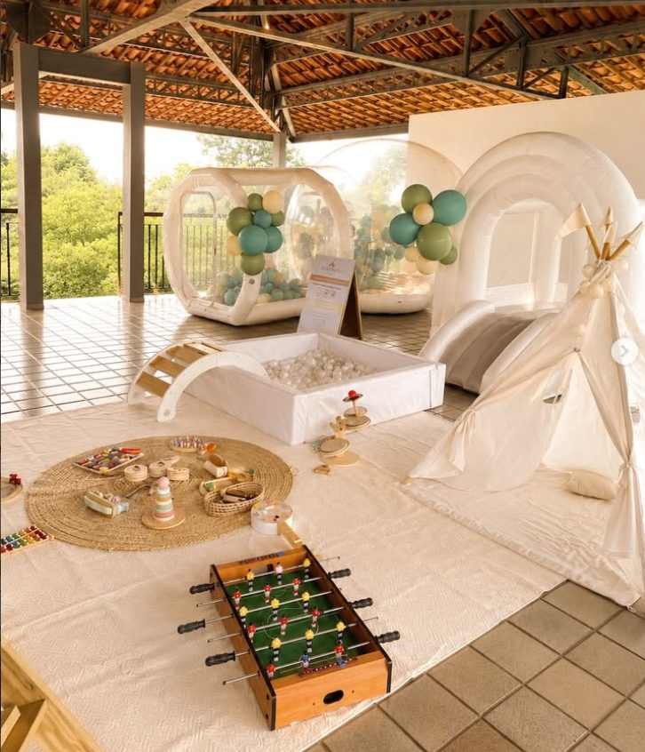 Espaço montado com Bubble House decorada com balões verdes, inflável arco-íris, cabana, piscina de bolinhas, pebolim e brinquedos de madeira